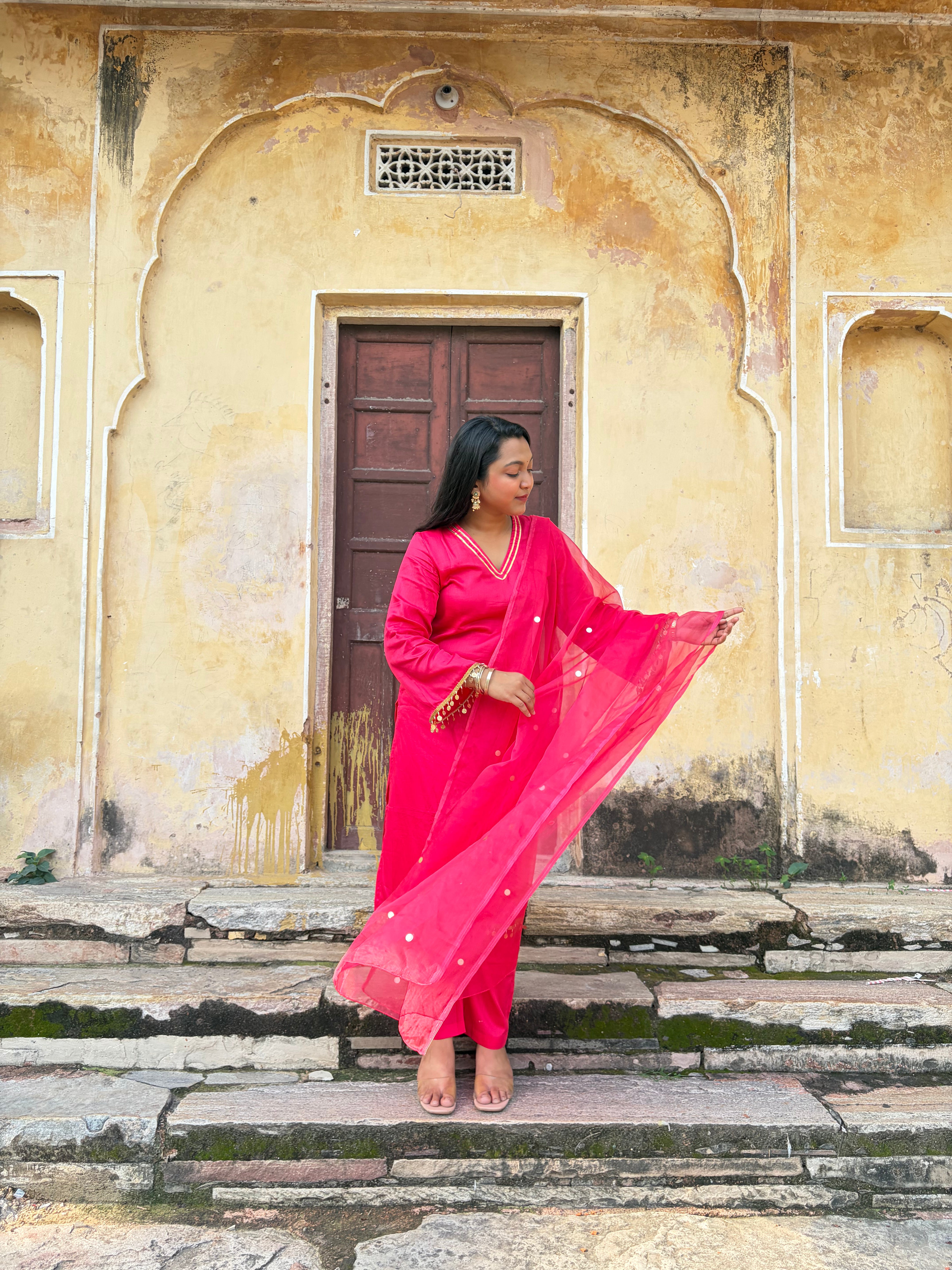 A woman wearing a bright pink traditional outfit with a matching sheer dupatta stands on stone steps in front of a weathered beige stone building with a brown door.