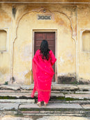 A woman viewed from behind wearing a bright pink traditional outfit with a matching dupatta, standing on stone steps in front of a weathered yellow-beige building with a wooden door.