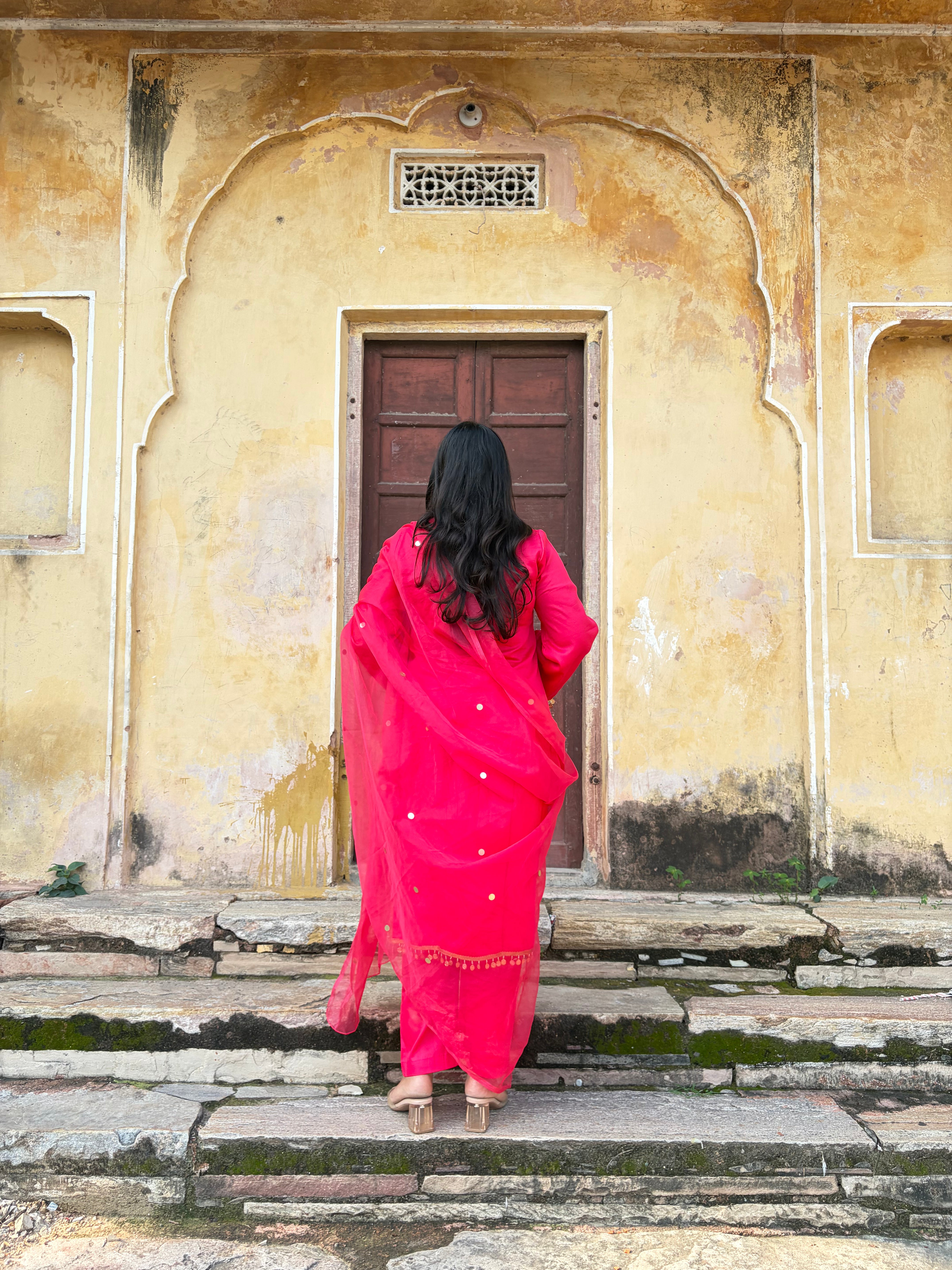A woman viewed from behind wearing a bright pink traditional outfit with a matching dupatta, standing on stone steps in front of a weathered yellow-beige building with a wooden door.
