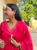 A woman wearing a bright pink traditional outfit with gold trim and matching sheer dupatta, standing outdoors near a building and trees.