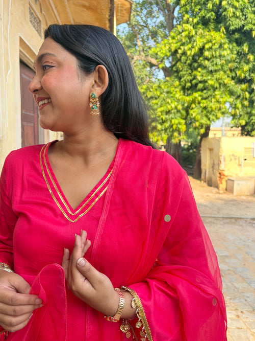 A woman wearing a bright pink traditional outfit with gold trim and matching sheer dupatta, standing outdoors near a building and trees.