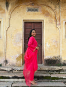 A woman wearing a bright pink traditional outfit with a matching dupatta stands on stone steps in front of a weathered beige wall with a dark wooden door.