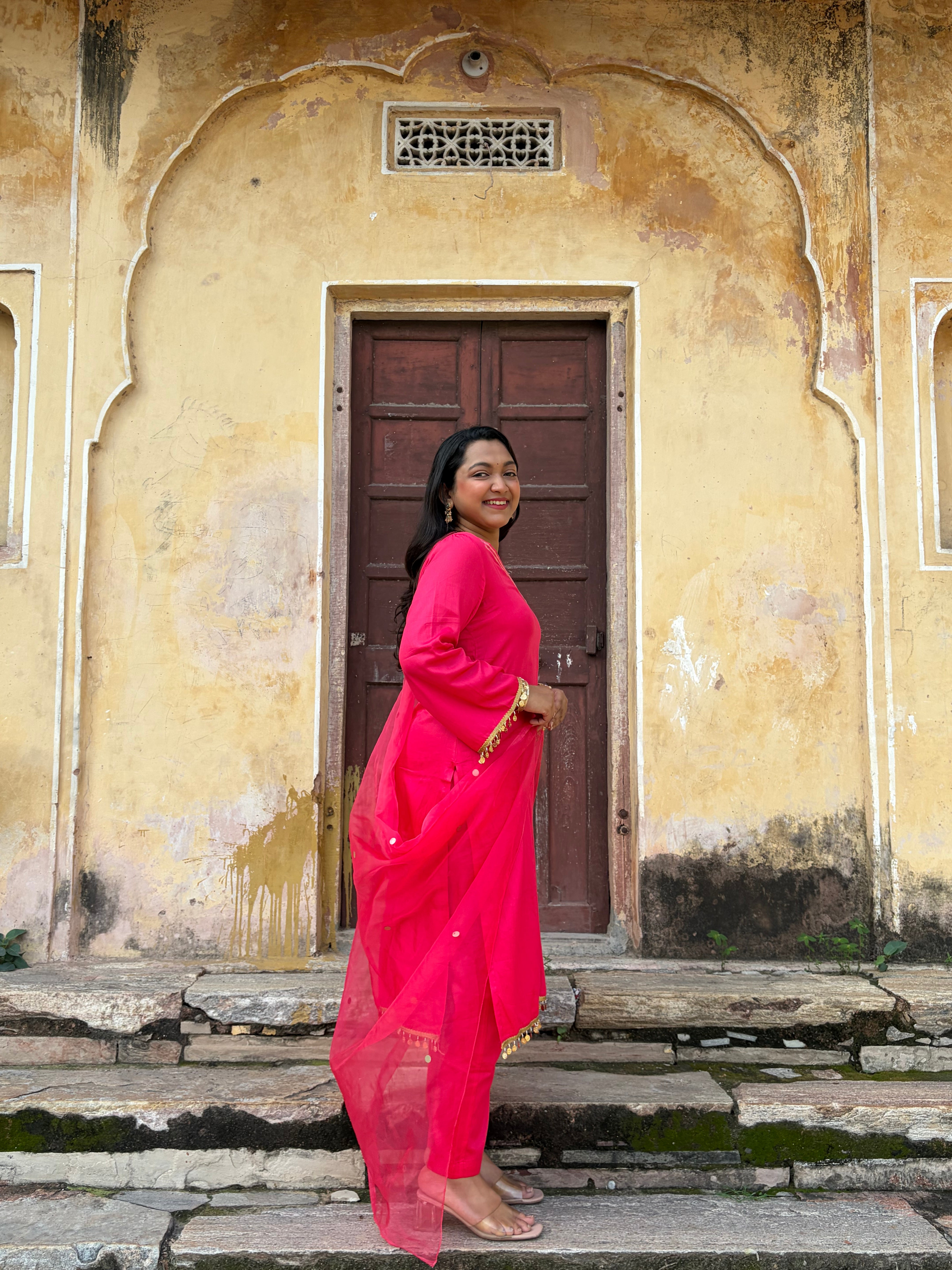 A woman wearing a bright pink traditional outfit with a matching dupatta stands on stone steps in front of a weathered beige wall with a dark wooden door.