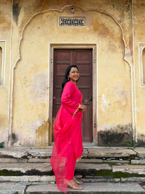 A woman wearing a bright pink traditional outfit with a matching dupatta stands on stone steps in front of a weathered beige wall with a dark wooden door.